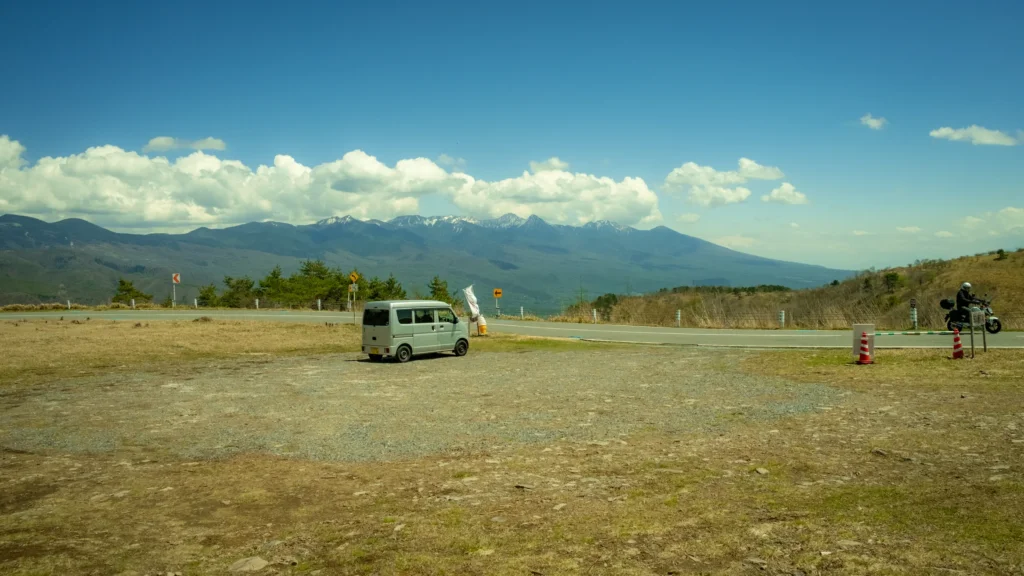 Parking en terre avec une camionnette et un motard face à la chaîne des Alpes japonaises sous de gros nuages