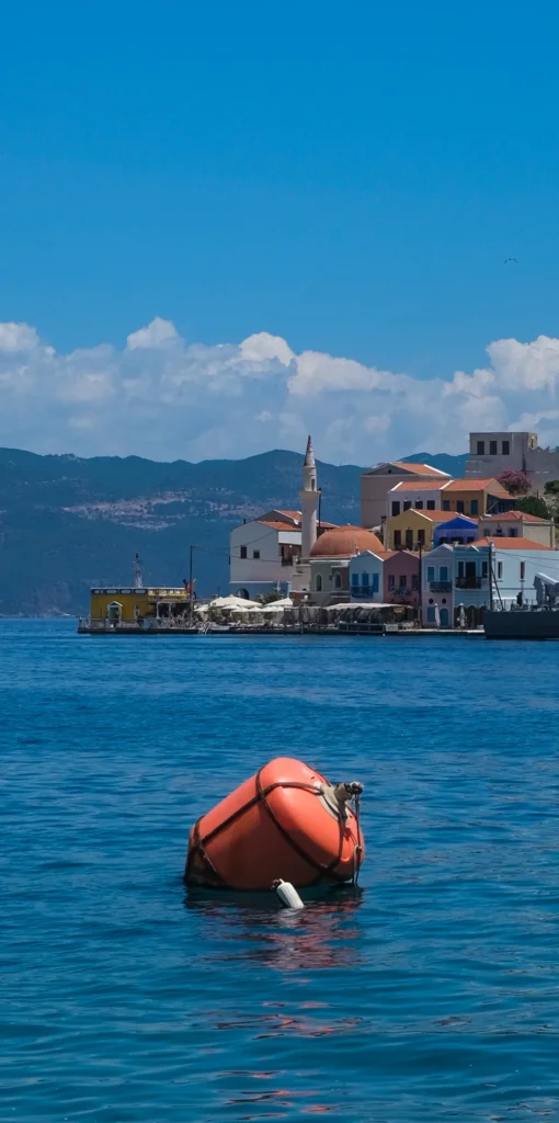 Grande bouée orange flottant sur une mer bleue face aux maisons colorées et à la mosquée du port de Yialos, Symi