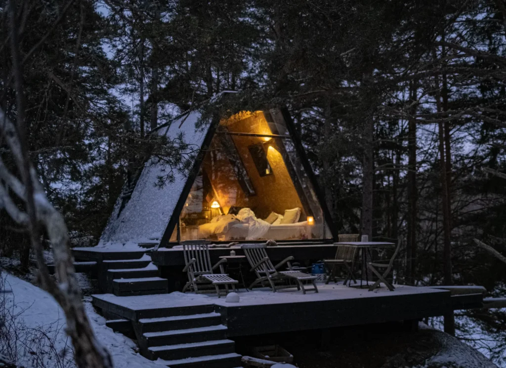 Cabane en A-frame aux parois vitrées, intérieur chaleureusement éclairé, terrasse en bois enneigée entourée de sapins