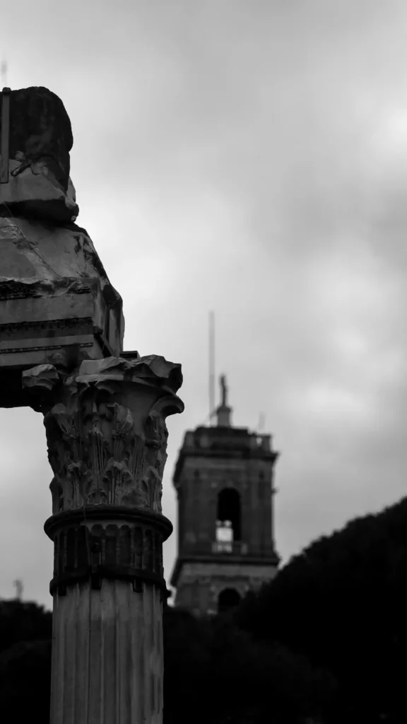 Photo en noir et blanc d'un chapiteau corinthien en ruine au premier plan, avec le clocher de Santa Francesca Romana en arriere-plan flou