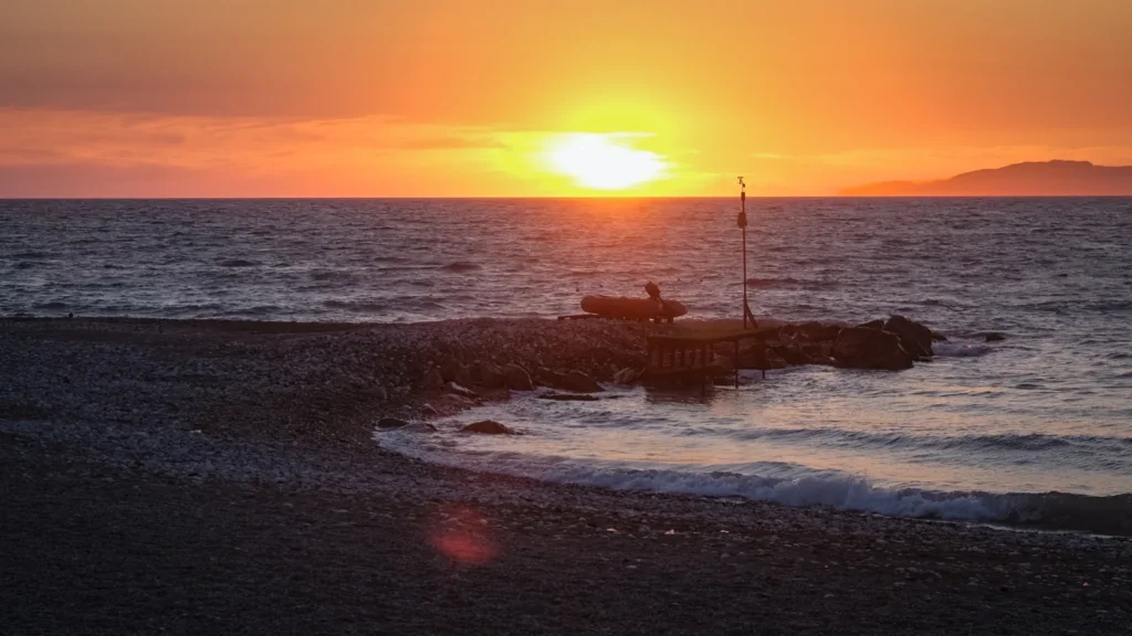 Coucher de soleil orange sur la mer, ponton et bateau pneumatique sur une plage de galets à Theologos, Kastellórizo