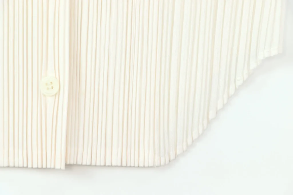Gros plan sur le bas plissé écru d'un gilet Issey Miyake Pleats Please, avec un bouton nacré au premier plan