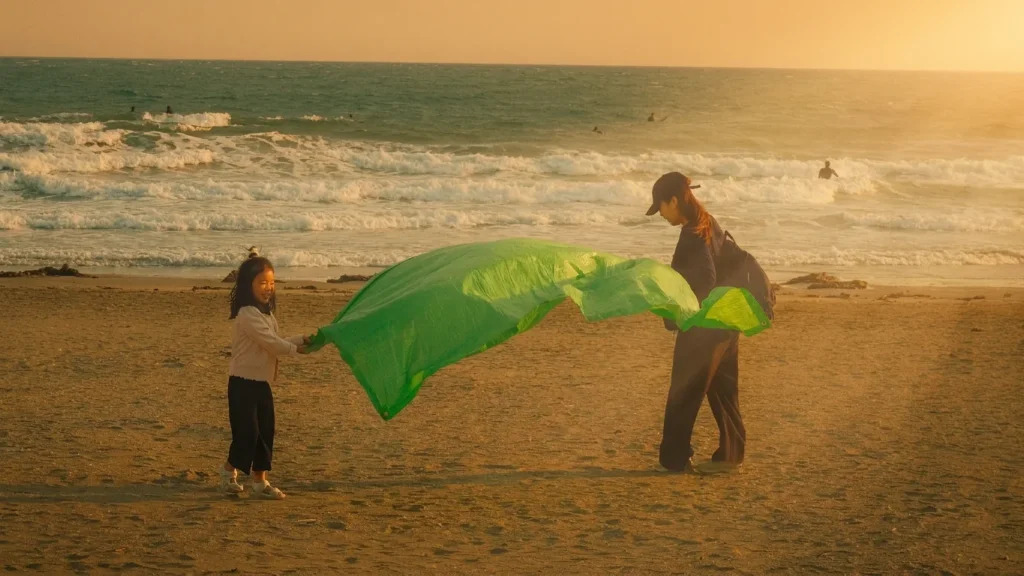 Enfant et adulte tenant une grande voile verte au vent sur une plage au coucher du soleil, vagues et surfeurs au fond