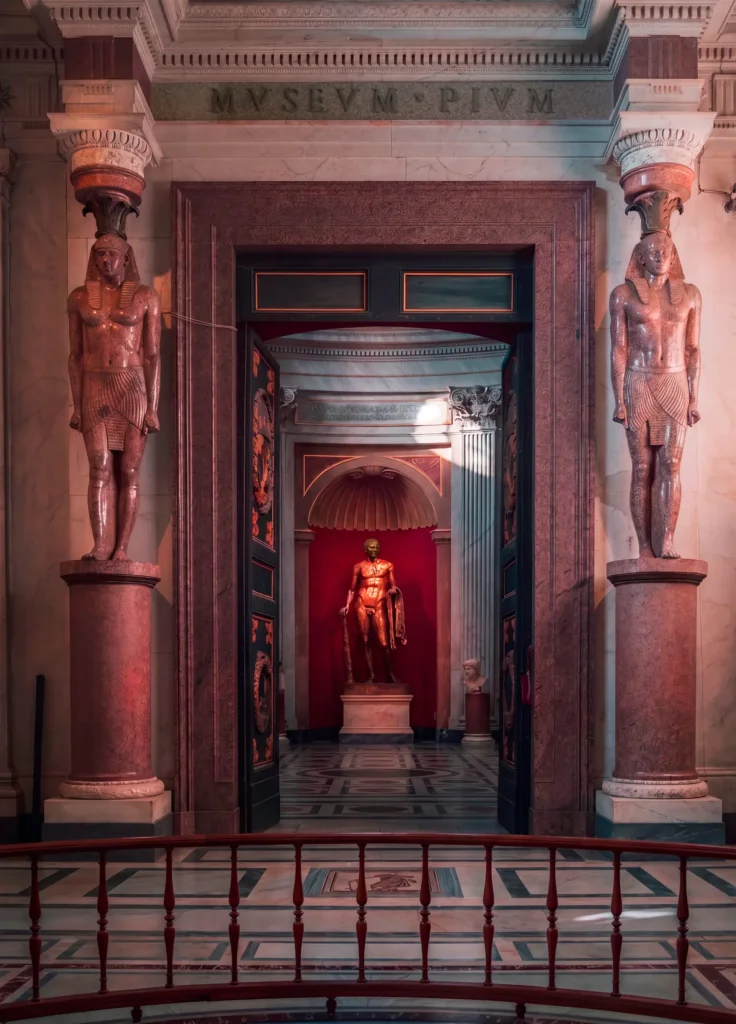 Vue en enfilade depuis un balcon vers l'entrée du Museo Pio-Clementino, encadrée de statues égyptisantes, statue de bronze doré en fond