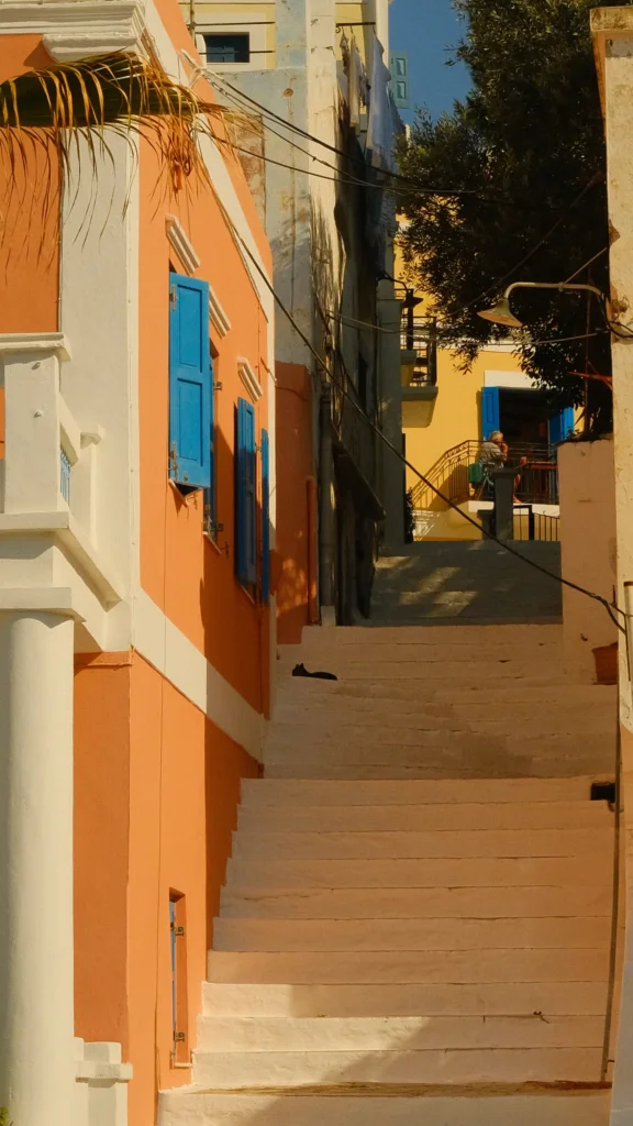 Escalier extérieur bordé de façades orange et jaune à volets bleus, dans la citadelle de Symi
