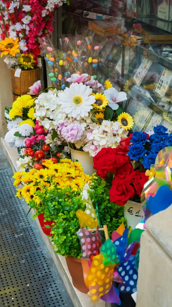 Étalage extérieur d'une boutique proposant des fleurs artificielles multicolores (roses, marguerites, tournesols) devant une vitrine de téléphonie