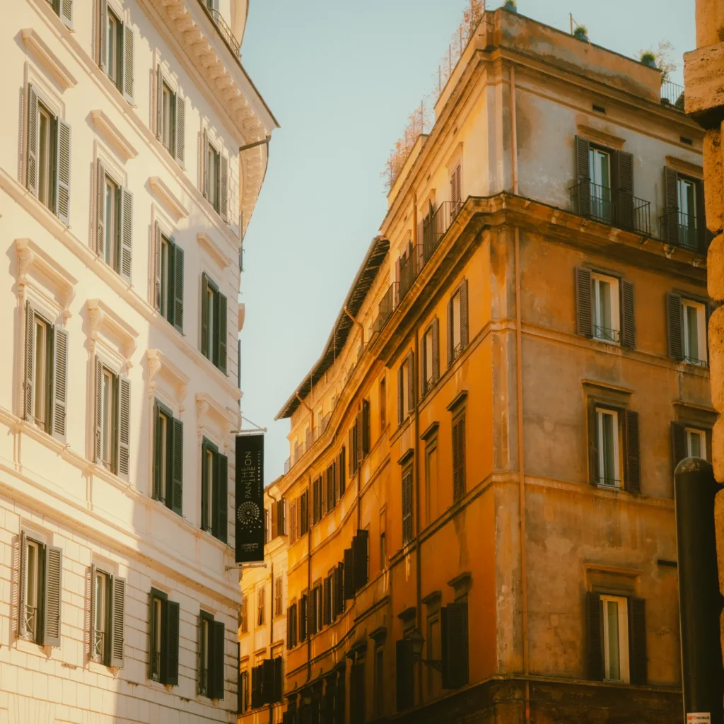 Vue en contre-plongée d'une ruelle romaine bordée d'immeubles blanc et ocre, enseigne verticale de l'hôtel Pantheon Iconic Rome