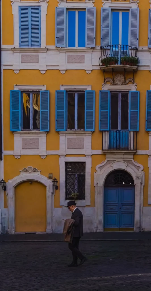 Homme au chapeau consultant son téléphone devant une façade jaune ornée de volets bleus et d'une porte numérotée 170, style romain