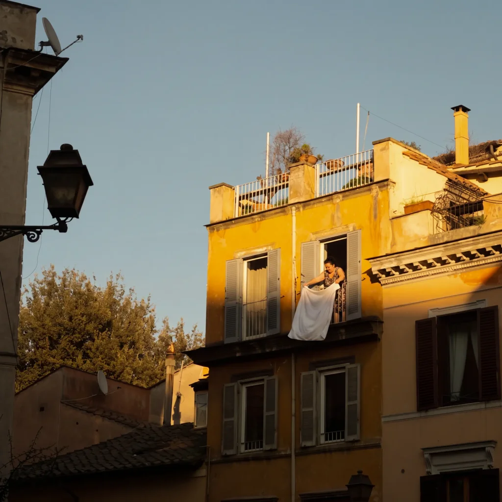 Femme secouant un drap blanc a la fenetre d'un immeuble jaune, lumiere doree de fin de journee, volets gris ouverts