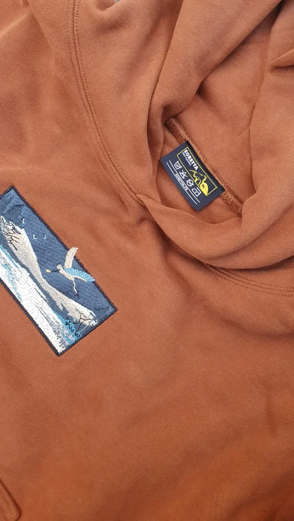 Gros plan sur l'étiquette Egretta et un patch brodé représentant une scène de plongeon, sur un hoodie orange brique