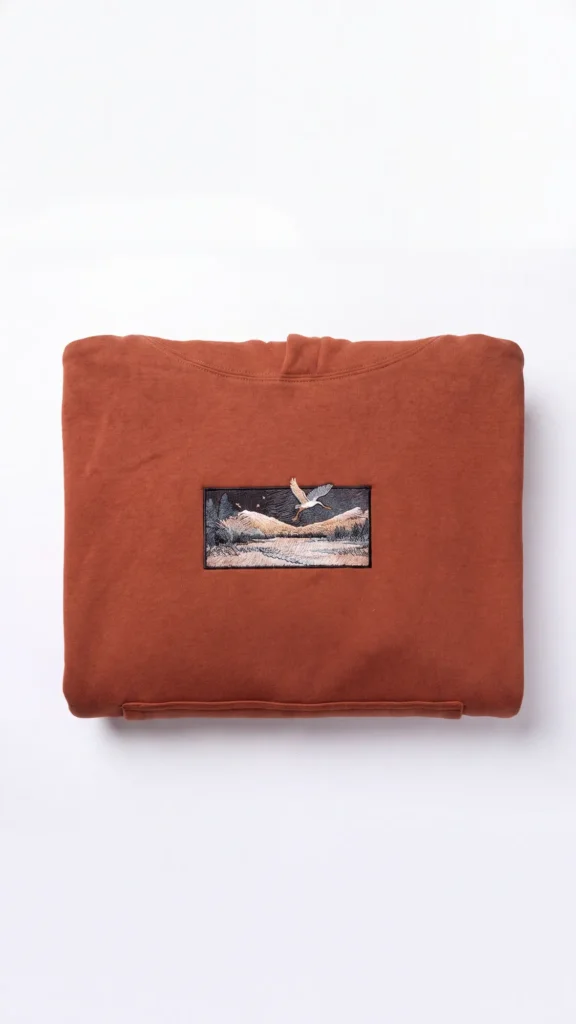Hoodie Egretta orange brique plié à plat, patch brodé aigrette et paysage de montagne