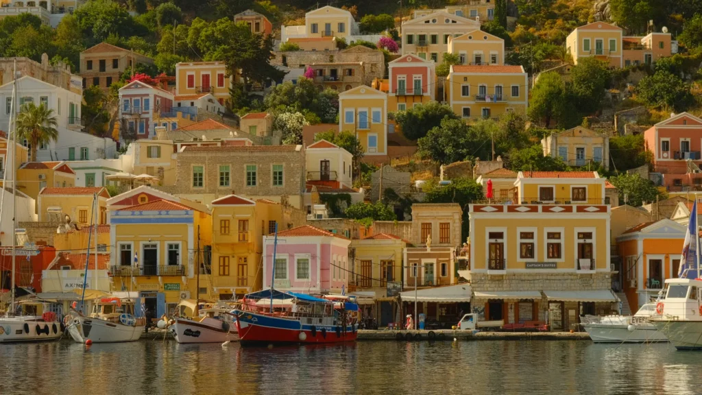 Gros plan sur les façades jaunes, roses et orange des maisons du port de Symi, bateaux amarrés au premier plan