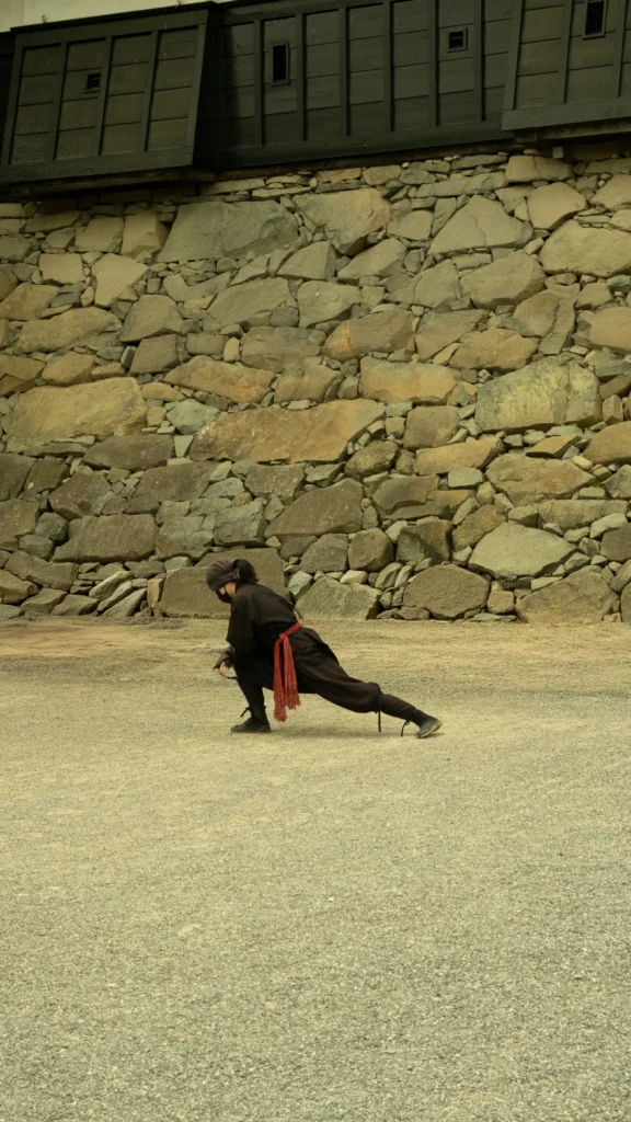 Figurant en costume de ninja noir et rouge, en fente au sol devant un mur de pierre du château de Matsumoto