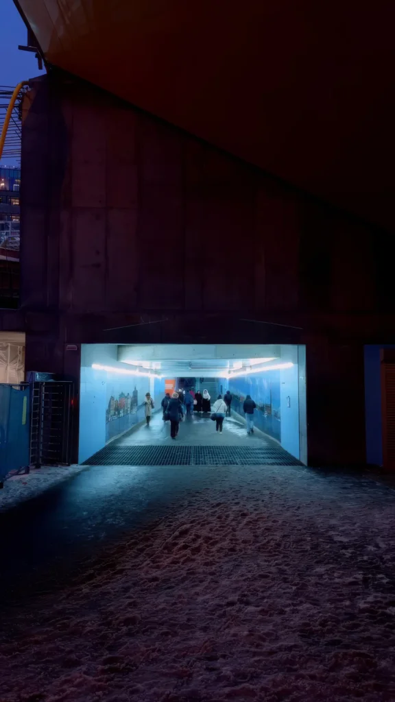Tunnel piéton éclairé en bleu la nuit, fresque représentant la skyline de la ville sur les parois, passants en mouvement