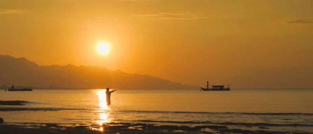 Silhouette d'un pêcheur dans l'eau au coucher de soleil, bateaux traditionnels et montagnes dans le nord de Bali