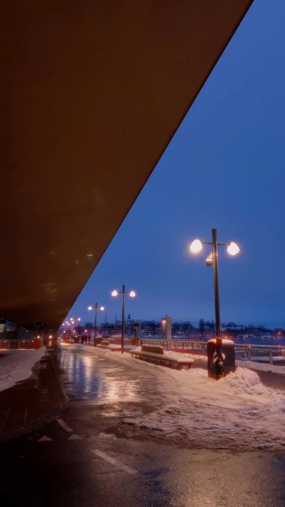 Promenade enneigée le long de l'eau à la tombée de la nuit, lampadaires allumés, vue sur la ville de l'autre côté du plan d'eau, sous l'auvent du nouveau terminal de Slussen