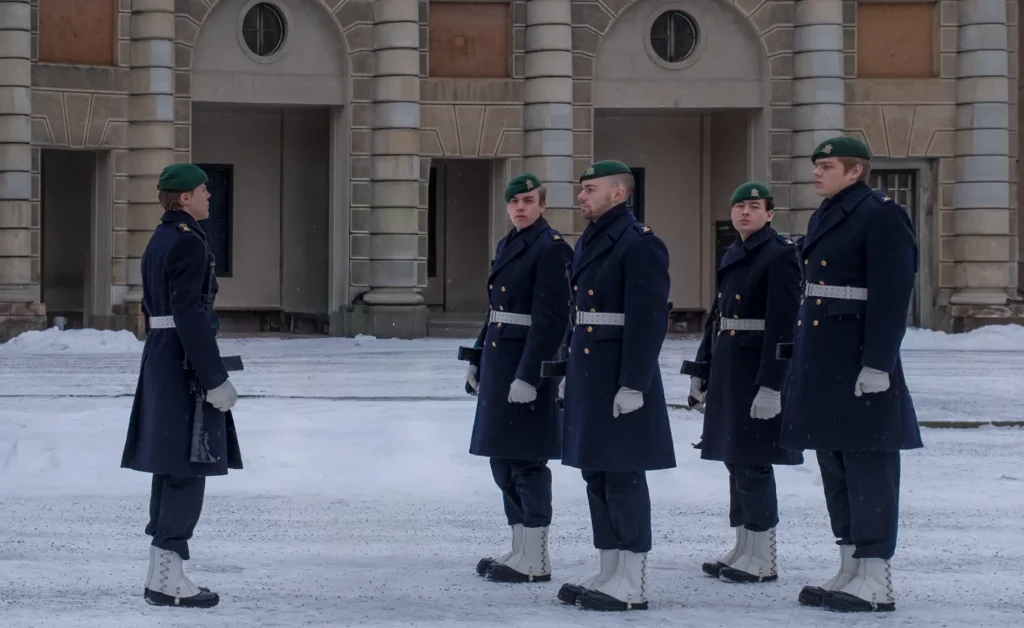 Cinq gardes royaux suédois en manteau bleu marine et béret vert alignés dans la cour enneigée devant les anciennes écuries du palais de Drottningholm