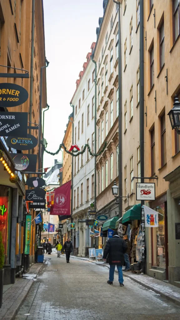 Ruelle pavée bordée de boutiques et enseignes colorées, décorations de Noël suspendues entre les façades, passants dans la Gamla Stan