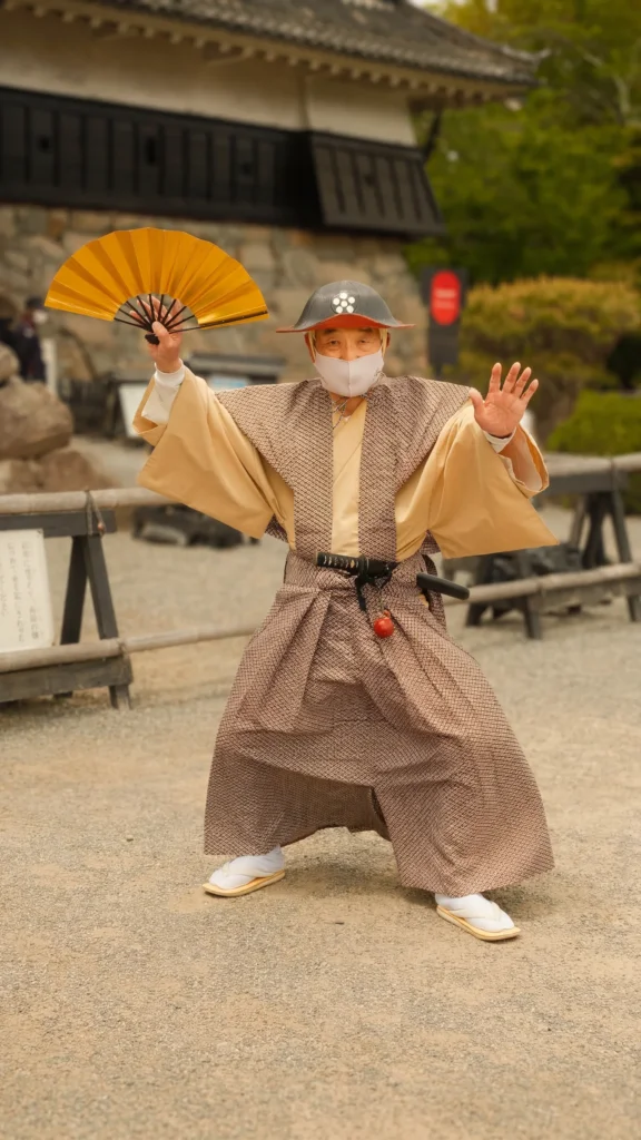 Homme en costume de samouraï brandissant un éventail doré dans la cour du château de Matsumoto