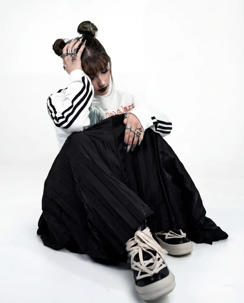 Modèle assise en sweat rayé blanc et noir et longue jupe plissée noire, bagues griffées, shooting Egretta en studio