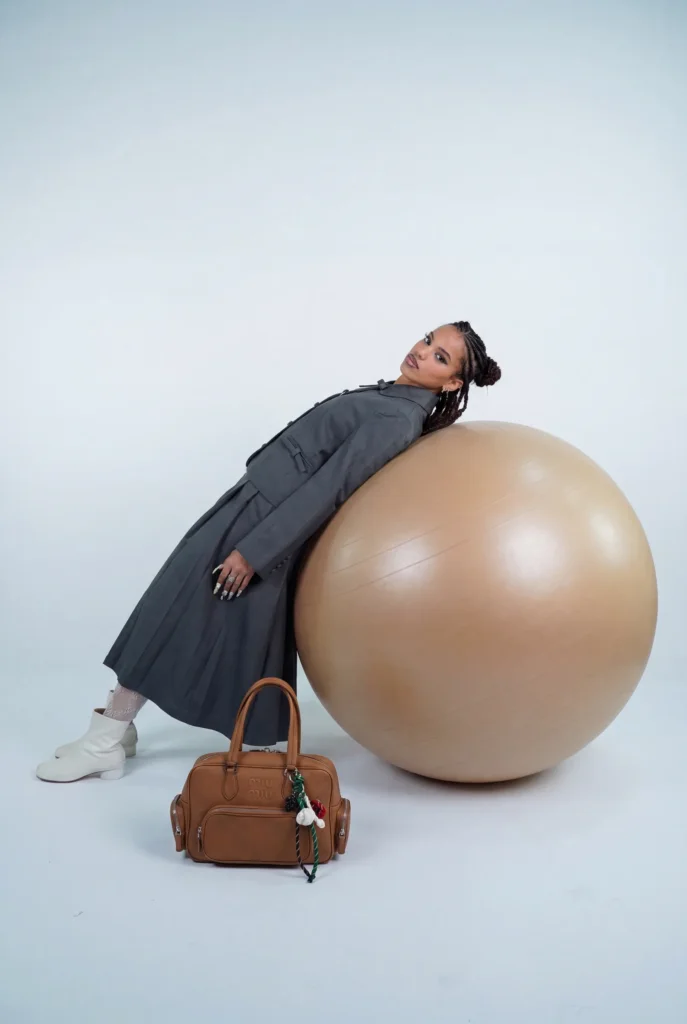 Portrait studio d'une modèle en trench gris appuyée sur un gros ballon, avec un sac Miu Miu au premier plan, shooting Egretta