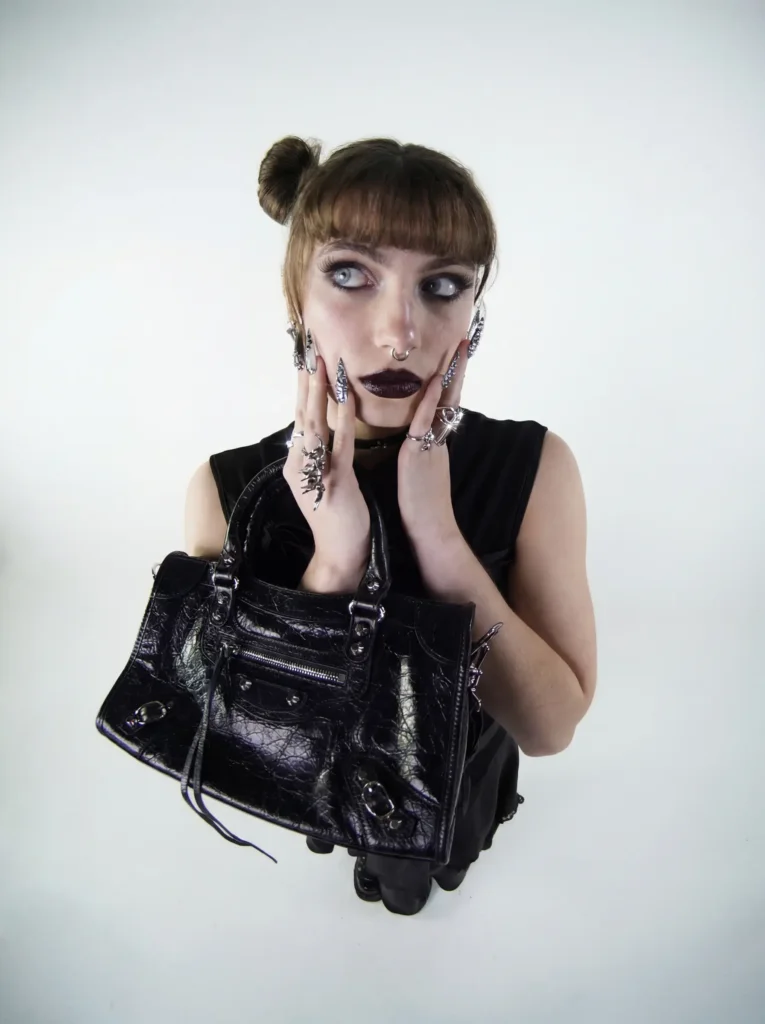 Portrait en plongée, femme tenant un sac à main noir texturé à double poignée, mains sur le visage, ongles argentés griffés, fond blanc