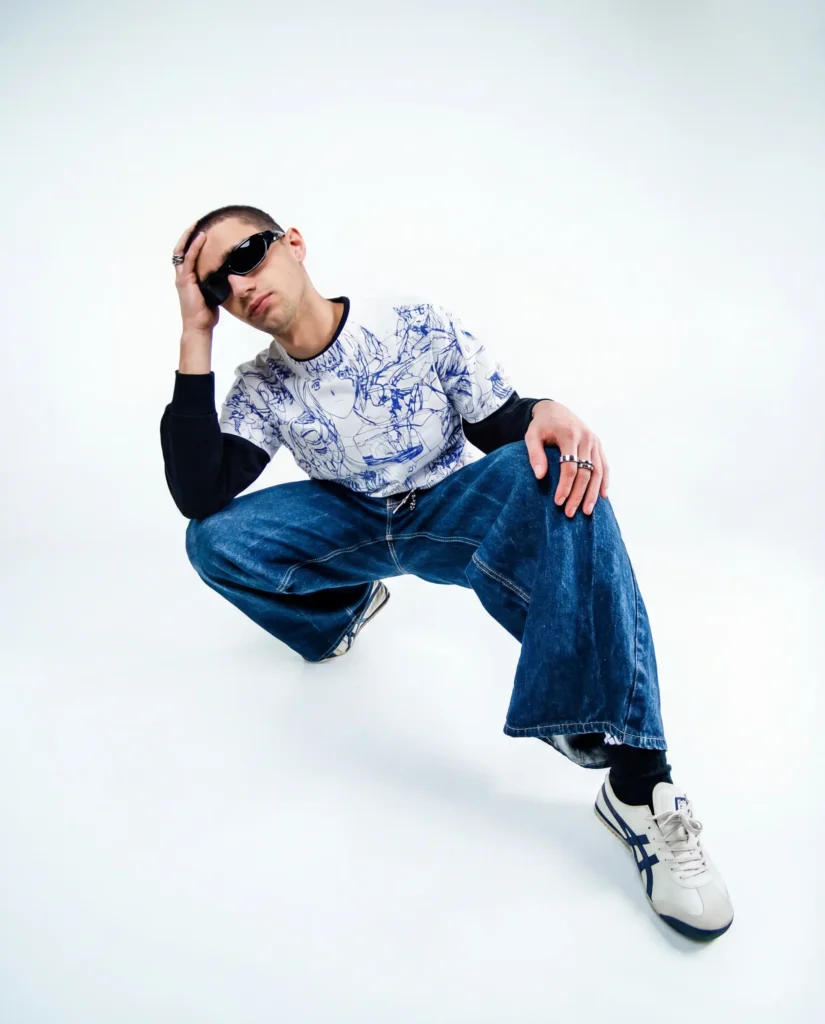 Homme accroupi en studio, sweat imprimé manga bleu et blanc, jean large délavé, lunettes de soleil ovales, sneakers blanches à bandes bleues