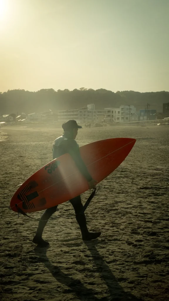 Surfeur portant sa planche rouge sur une plage de sable à contre-jour, bâtiments au loin