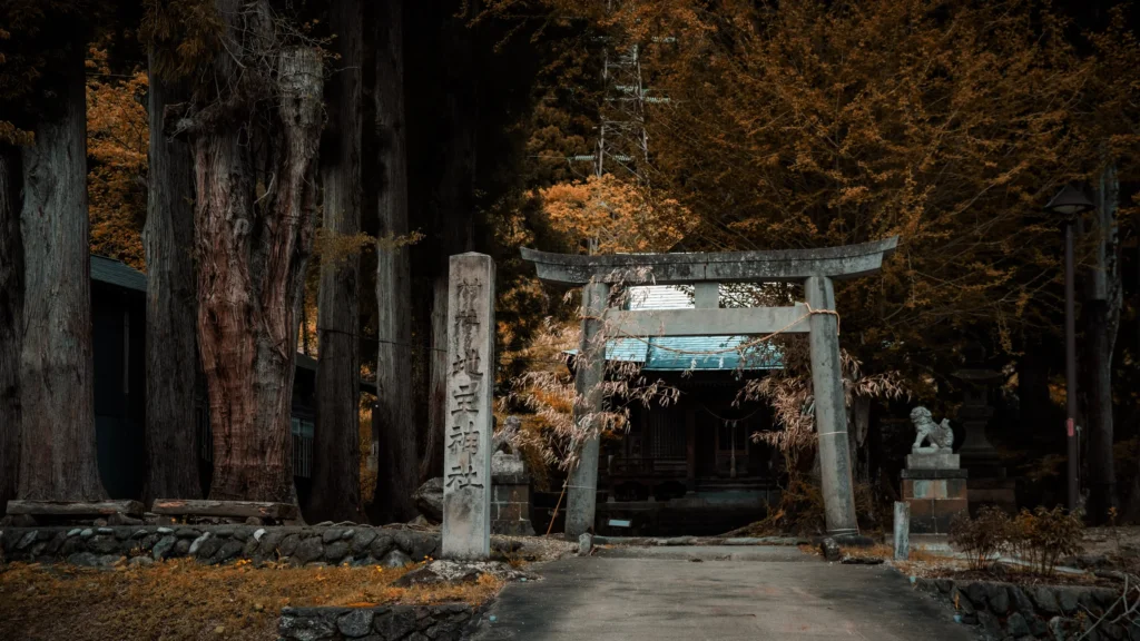 Torii de pierre et stèle devant un sanctuaire au milieu de grands cèdres et de feuillages cuivrés