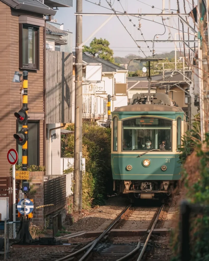 Tramway Enoden vert arrivant sur une voie bordée de maisons et de poteaux de passage à niveau jaunes et noirs