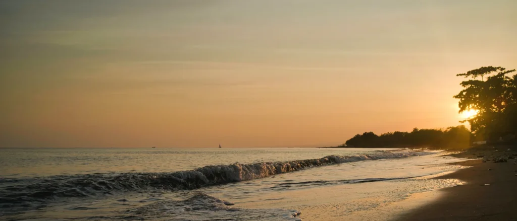 Vague s'échouant sur une plage de sable noir au coucher de soleil, silhouette d'arbres et voilier au loin, Seririt, Bali