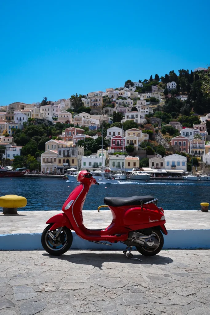 Scooter Vespa rouge garé sur le quai face aux maisons colorées et bateaux du port de Symi