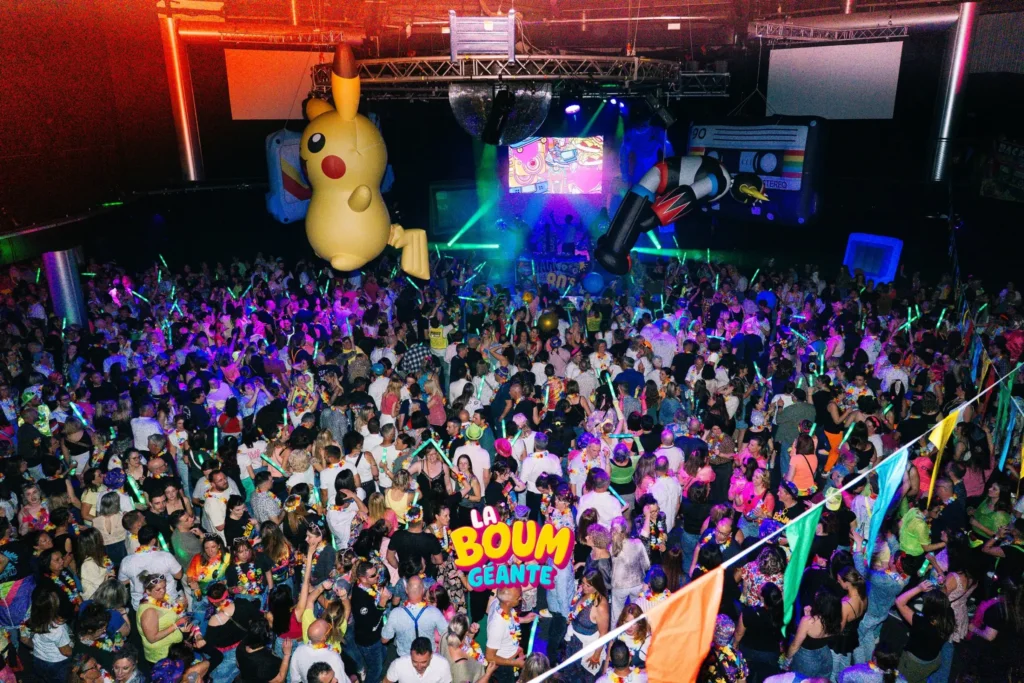 Foule dense dansant avec des bâtons lumineux sous un Pikachu gonflable géant suspendu, lors de la soirée La Boum Géante à Clermont-Ferrand